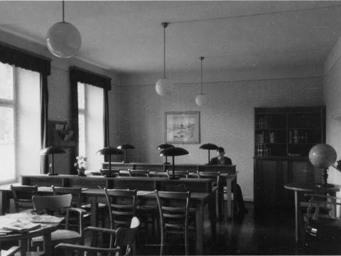 Auf der Schwarz-Weiß-Fotografie sieht man einen Raum mit zwei Tischreihen und Stühlen. Jeder Arbeitsplatz ist mit einer Lampe ausgestattet. An einem sitzt eine Person. In der hinteren rechten Ecke steht ein Bücherregal.