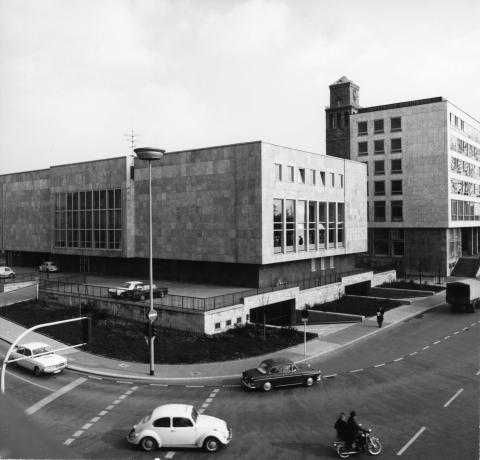 Die Schwarz-Weiß-Fotografie zeigt das Gebäude der Stadtbücherei am Rathausmarkt mit dem Rathausturm im Hintergrund. Auf der Straße vor dem Gebäude fahren Autos und ein Motorrad.