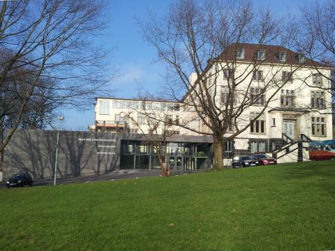 Zu sehen ist das sanierte historische Gebäude der alten Augenklinik mit einem modernen Anbau. Im Vordergrund des Bildes sind eine grüne Wiese und Bäume am Straßenrand.