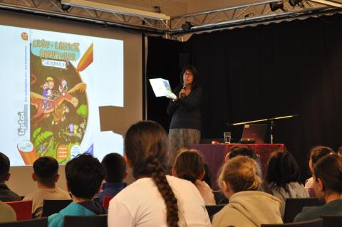 Auf dem Bild sieht man die Autorin Annette Neubauer auf der Bühne. Sie spricht in ein Mikrofon und stellt ihr Buch vor. Das Cover des Buches wird auf einer Leinwand angezeigt. Im Vordergrund sieht man viele Kinder von hinten.