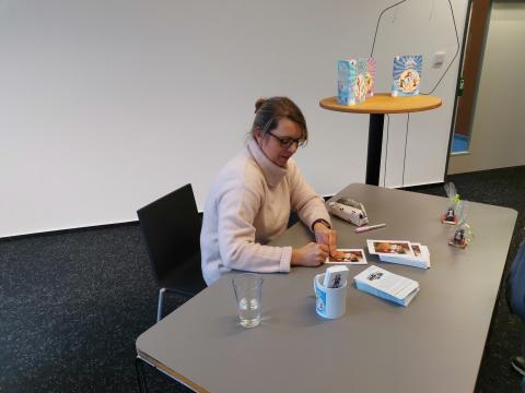 Auf dem Bild sieht man die Autorin Angelika Hesse. Sie sitzt an einem Tisch und unterschreibt Autogrammkarten. Im Hintergrund werden ihre Bücher auf einem Stehtisch präsentiert.