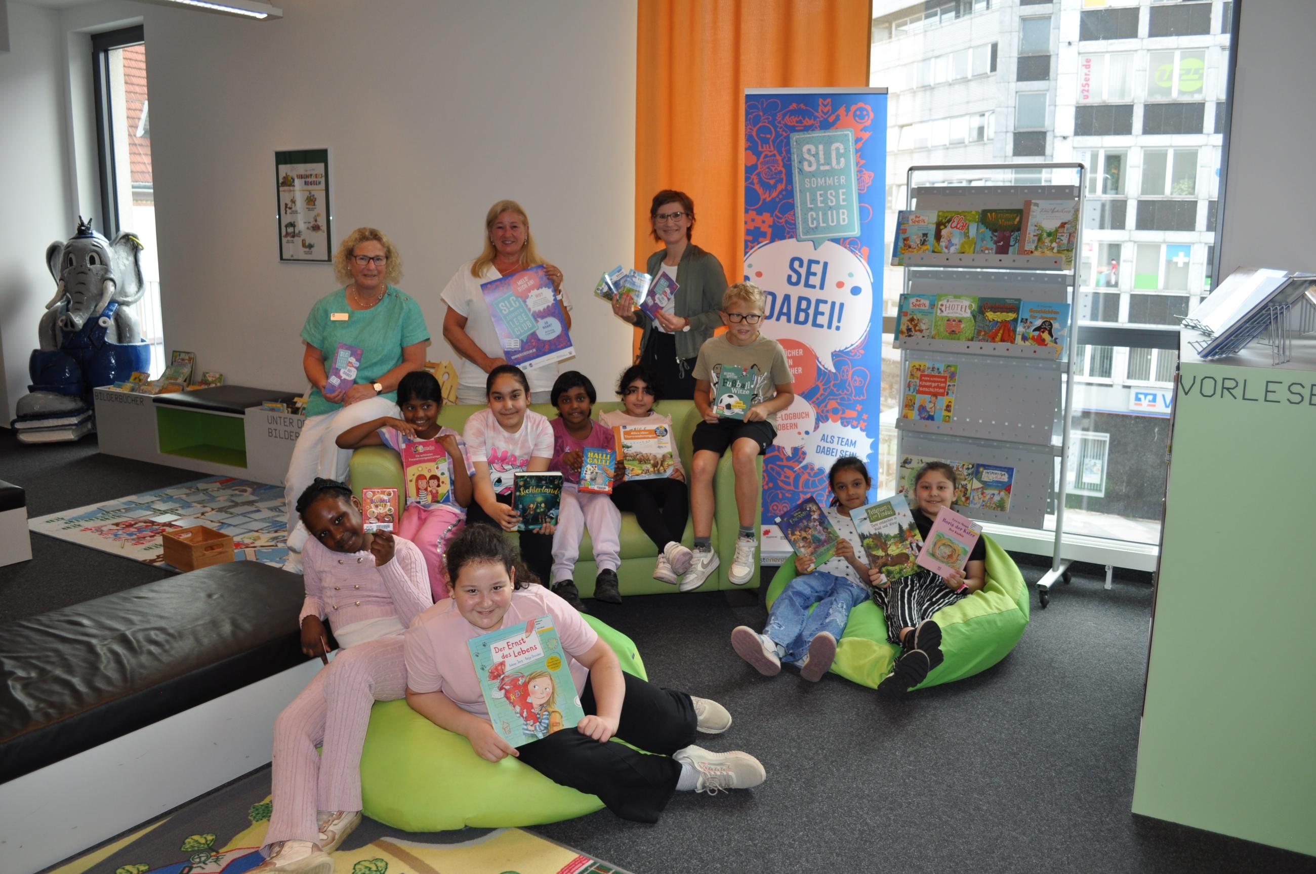 Ankündigung des SommerLeseClubs in der Stadtbibliothek Mülheim an der Ruhr Vom Team des MedienHauses sind Elke Hoffmann (stellvertretende Bibliotheksleiterin) und Luciana Turra-Fabriczius (Lektorin Jugendliteratur) sowie Simone Kastner-Zens (Kommunalmanagerin Westenergie) mit Schüler*innen der Pestalozzi-Schule zu sehen.