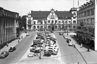 <p><span>Der Synagogenplatz, vormals Viktoriaplatz, in den 1950er-Jahren als Parkfläche genutzt, links das damalige Sparkassengebäude, Fotografie, Medienkompetenzzentrum</span></p>