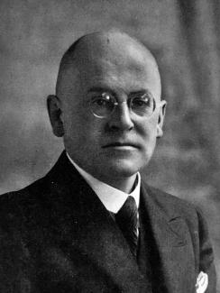 Porträt von Dr. Alfred Schmidt. Er hat eine Glatze. Er trägt eine Brille mit kleinen runden Gläsern und einen Anzug mit Krawatte und Einstecktuch.