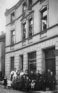 Das Wohnhaus der Familie Wolf im Jahr 1916, Scharpenberg 42. Vor dem Haus stehen Familienmitglieder, andere Bewohner*innen des Hauses und Bedienstete. Johanna und Ferdinand Wolf gucken aus den mittleren Fenstern der oberen Etage.