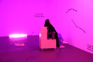 <p><span>Das Foto zeigt eine Person in der installativen Arbeit "when your identity runs away/how far does your identity run?" von Maria Reneé Morales Garcia. Sie sitzt auf einem Stuhl, den die Künstlerin wie auch die Wände und den Boden beschrieben hat.</span></p>