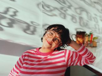 <p><span>Die Künstler*in Maria Reneé Morales Garcia blickt auf diesem Foto lächelnd in die Kamera. Von links fällt Licht durch ein Fenster auf sie.</span></p>