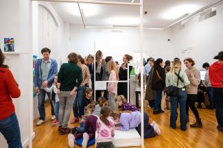 Viele Menschen, Erwachsene und Kinder, tummeln sich in einem Ausstellungsraum, unterhalten sich und schauen sich die Kunstwerke an.