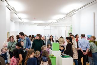 Viele Menschen, Erwachsene und Kinder, tummeln sich in einem Ausstellungsraum, unterhalten sich und schauen sich die Kunstwerke an.