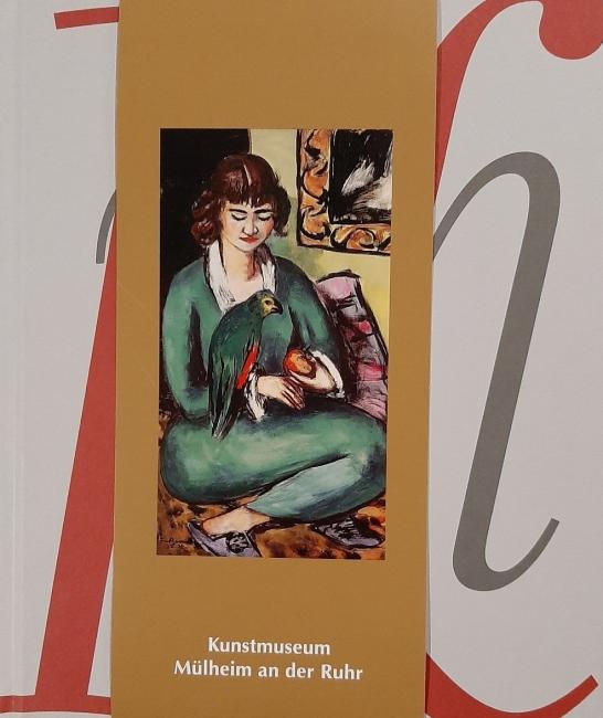 Das Bild zeigt das Cover des Sammlungskataloges - eine goldene Banderole mit Max Beckmanns Gemälde "Quappi mit Papagei" umschlingt ein graues Hardcover-Buch mit den verschlungenen roten und dunkelgrauen Buchstaben k, m und h.