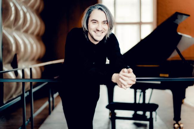 Das Bild zeigt den Pianisten Alexander Gadjiev mit einem Flügel im Hintergrund. Er tritt am 17. Januar 2025 als Solist in der Stadthalle bei den Sinfoniekonzerten auf.