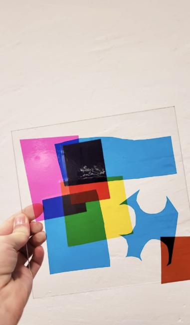 Das Foto zeigt eine Hand, die eine Plexiglasscheibe hält, die mit bunten, durchsichtigen Formen versehen ist. 