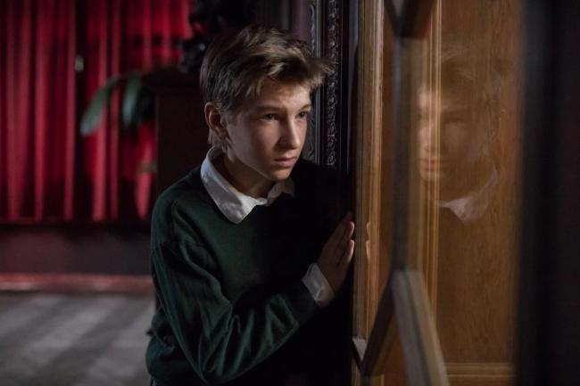 Ein Junge schaut vorsichtig aus einem Fenster. Im Hintergrund ist ein roter Vorhang.