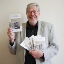 Porträt eines Mannes mit grauen Haaren, Dreitagebart und Brille, welcher mit Mülheimer Geschichtsheften posiert, die er vor sich in die Kamera hält. Er trägt ein schwarzes Hemd und ein hellgraues Sakko.