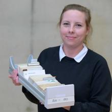 Porträt einer Frau mit zusammengebundenen, dunkelblonden Haaren, die die Schublade eines Zettelkatalogs in die Kamera hält. Sie trägt einen schwarzen Pullover, unter dem ein weißer Hemdkragen hervorragt.