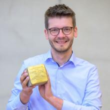 Porträt eines Mannes mit Kurzhaarfrisur und runder Brille, der einen Stolperstein in die Kamera hält. Er trägt ein hellblaues Hemd.