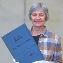 Porträt einer Frau mir grauer Kurzhaarfrisur, die eine Akte der Bürgermeisterei Broich in die Kamera hält. Der Aktendeckel ist dunkelblau. Die Frau trägt ein grau-, weiß- und beigefarbendkariertes Hemd, darunter ein schwarzes Oberteil und weiße Handschuhe