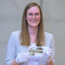 Porträt einer Frau mit dunkelblonden Haaren und runder Brille, die eine Postkarte mit Motiven der Stadt Mülheim in die Kamera hält. Sie trägt ein weißes Oberteil unter einem beigefarbenen Blazer und weiße Stoffhandschuhe.