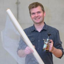 Porträt eines Mannes mit Kurzhaarfrisur, der in der einen Hand eine Rolle mit dünnem Stoff und in der anderen Hand ein metallenes Gefäß. Er trägt ein dunkelgraues Hemd.