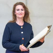 Porträt einer dunkelhaarigen Frau, die ein aufgeschlagenes großes Buch in den Händen hält. Sie trägt eine dunkelblaue Strickjacke mit goldenen Knöpfen.