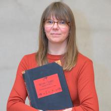 Porträt einer Frau mit schulterlangen, dunkelblonden Haaren und Brille. Sie trägt ein rotes Oberteil und hält ein schwarzes großes Buch in den Armen. Auf dem Buchdeckel klebt ein ein roter Aufkleber mit schwarzer Schrift.