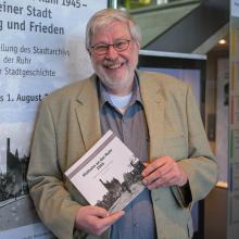 Dr. Stefan Pätzold hält die neue Publikation des Stadtarchivs "Mülheim an der Ruhr 1945" in der Hand und lächelt in die Kamera.